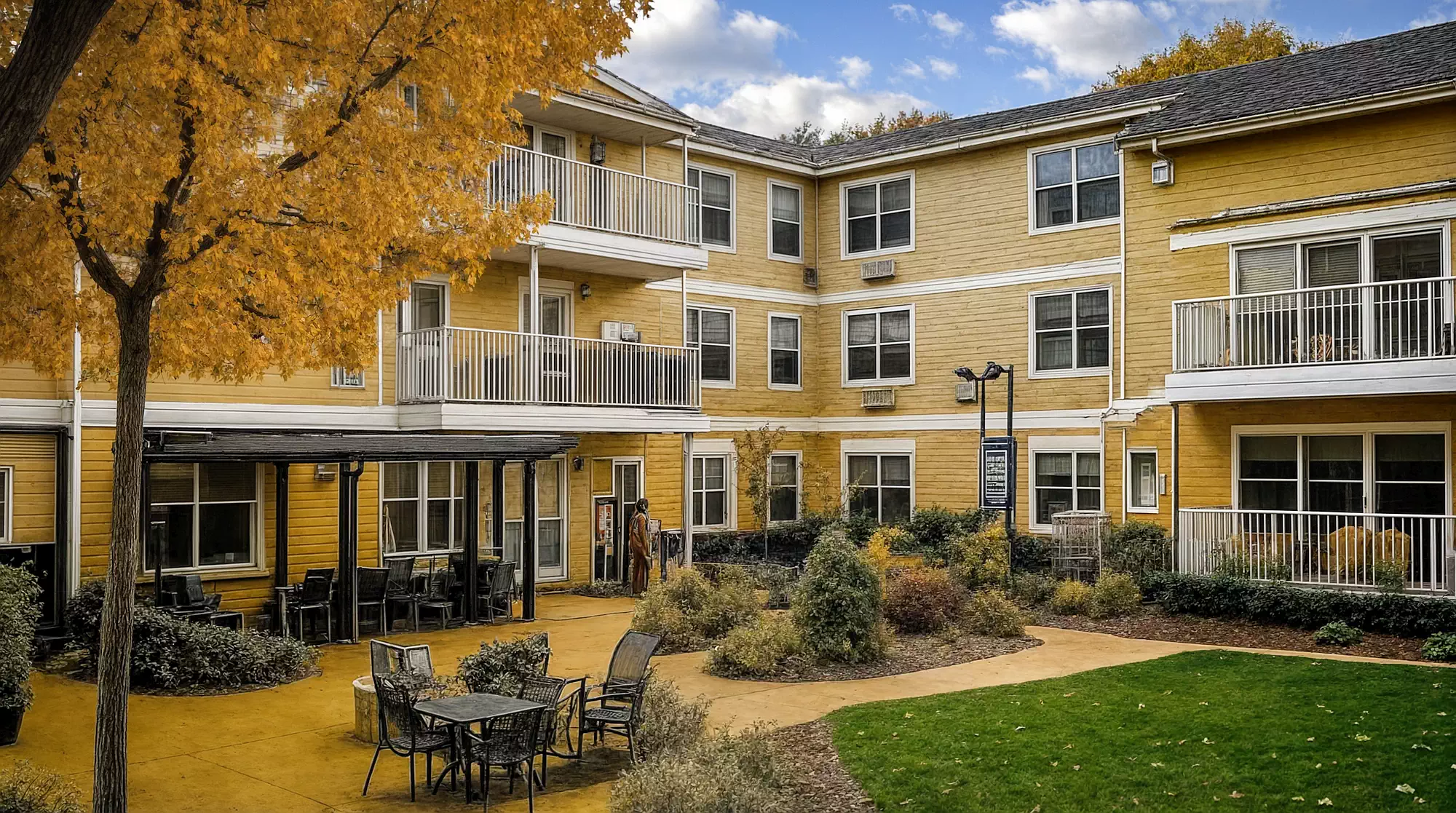 A three-story yellow building with balconies, this senior living facility is located around a courtyard featuring tables, chairs, and autumn foliage.
