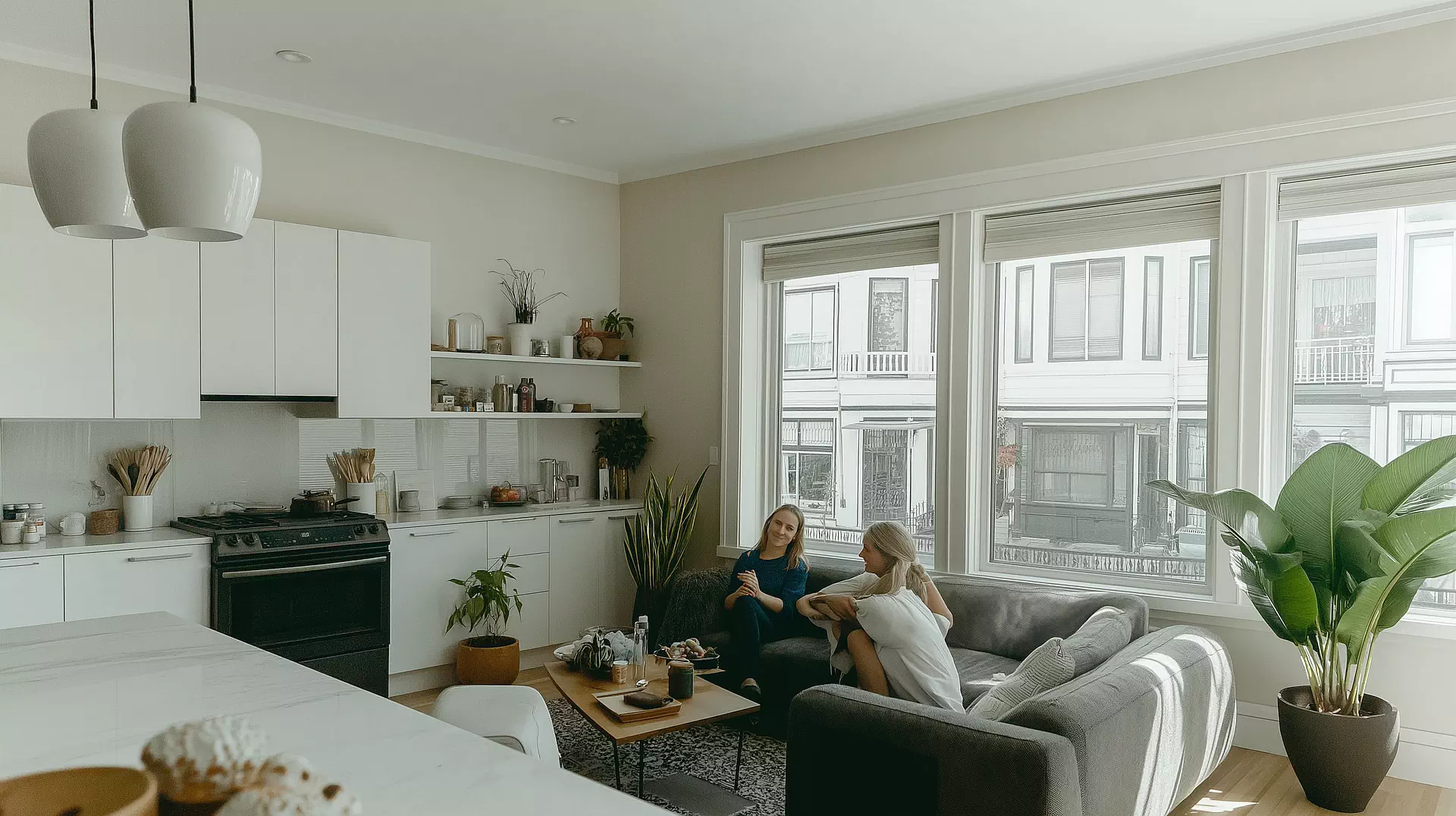 Two people sit and converse on a couch in a modern kitchen-living room, surrounded by potted plants and large windows, discussing the new Airbnb features.